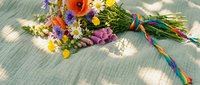 Colorful wildflower bouquet with poppies, daisies and purple blooms tied with rainbow ribbon on textured fabric surface for spring celebrations.