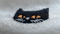 Black cat face embroidered on white fabric with orange eyes and whisker details, close-up macro view showing textile texture and stitching pattern.