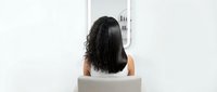 Young woman with curly dark hair sitting on white chair in minimalist modern interior with clean lines and bright natural lighting for wellness concepts.
