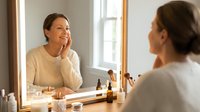 Young woman applying skincare routine looking at mirror reflection in bright bathroom with natural light and cosmetic products on vanity table.
