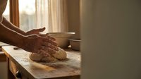 Senior baker hands kneading fresh bread dough on wooden counter in warm kitchen with natural sunlight streaming through window creating artisan bakery atmosphere.