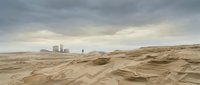 Industrial complex with smokestacks visible across vast, undulating sand dunes under overcast sky, suitable for environmental and industrial themes.