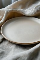 Minimalist ceramic plate on natural linen tablecloth with soft shadows and gentle folds, creating serene dining atmosphere in neutral beige tones.