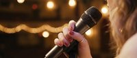 Young woman singing into microphone at evening event with warm bokeh lights creating atmospheric entertainment venue background for live music performances.