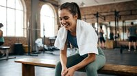 Happy young Hispanic woman resting on wooden bench after workout in modern gym with towel around neck smiling at camera during fitness break.