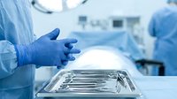 Surgeon in blue gloves reaching for sterile surgical instruments on metal tray in modern operating room during medical procedure preparation.