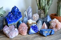 Collection of healing crystals and mineral specimens including azurite, rose quartz, clear quartz points and clusters arranged on wooden surface with potted plants.