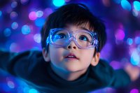 Young Asian boy wearing protective glasses gazes upward with wonder against vibrant blue and purple bokeh lights background, creating magical atmosphere for science and discovery themes.