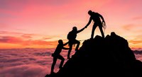 Silhouettes of hikers helping each other climb rocky mountain peak during dramatic sunset with pink and orange sky above clouds for teamwork concepts.