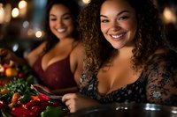 Joyful curly-haired mixed race woman smiling while preparing food with friend at evening dinner party. Warm bokeh lighting creates intimate atmosphere.