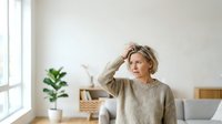Middle-aged woman with blonde hair wearing a beige sweater, touching her head thoughtfully in a bright, minimalist home office with potted plant and natural window light.