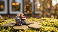 Two adorable ceramic figurines of elderly couple sitting together on wooden log slices in autumn garden with warm bokeh lights creating romantic atmosphere.