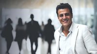 Confident Middle Eastern businessman in white suit smiling in corporate environment with blurred silhouettes of people in background.