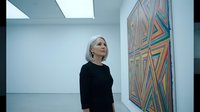 Middle-aged Caucasian woman in black outfit contemplating colorful geometric abstract artwork in modern white gallery space with natural lighting.