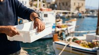 Mature male fisherman with dark skin holding white styrofoam container at harbor with moored boats, suitable for seafood, fishing industry, and maritime commerce content.