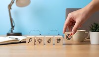 Hand placing wooden blocks with business icons including target, lightbulb, rocket and gears on desk representing strategic planning and goal achievement.