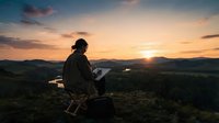 Silhouetted male freelancer working on laptop at sunset from mountain hilltop overlooking valley landscape, remote work and digital nomad lifestyle concept.
