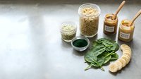 Superfood ingredients for healthy smoothies including spirulina powder, protein powder, nut butter jars, fresh spinach leaves and sliced banana on marble counter.