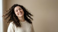 Happy young woman with curly hair laughing joyfully in cozy cream sweater against neutral background for lifestyle and wellness content.