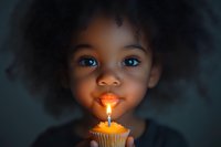 Young African American girl with bright blue eyes and natural curly hair smiling at glowing birthday cupcake with single lit candle against dark background.
