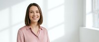 Young professional Caucasian businesswoman with brown bob haircut wearing pink blouse smiling confidently in modern bright office interior.