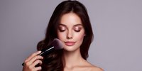 Young woman with closed eyes and pink makeup having powder brush applied to face against purple background. Perfect for beauty tutorials.