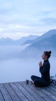 Woman practicing yoga meditation on wooden deck overlooking misty mountain landscape at dawn with prayer hands pose and peaceful atmosphere.