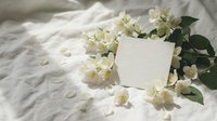 Blank cream card surrounded by white jasmine flowers and green leaves on soft linen fabric, suitable for wedding invitations, greeting cards, and stationery design projects.