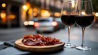 Two glasses of red wine with charcuterie board featuring cured meats on wooden plate. Evening city street background with warm bokeh lights creates romantic dining atmosphere.