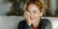 Young blonde woman smiling warmly while resting chin on hand, wearing cozy gray sweater in comfortable home setting for lifestyle and wellness content.