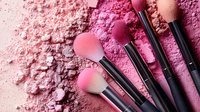 Makeup brushes arranged on pink crushed cosmetic powder background with scattered blush and eyeshadow pigments creating artistic texture and composition.