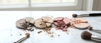 Crushed makeup compacts with scattered powder and brushes on a white surface, showcasing a neutral and rosy beauty palette.