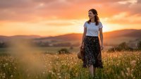 Woman with long brown hair, Caucasian ethnicity, wearing casual white t-shirt and floral skirt, standing in wildflower meadow at golden hour sunset. Suitable for lifestyle, travel, and wellness content.