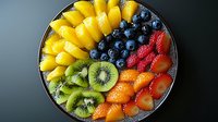 Fresh colorful fruit platter with mango slices, blueberries, raspberries, kiwi, and mandarin segments arranged in vibrant rainbow pattern on dark plate.