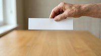 Senior male hand inserting blank white ballot paper into wooden box during voting process, election concept for civic participation and democratic rights.
