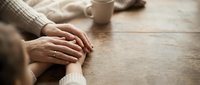 Caring hands gently holding together on wooden table with warm coffee cup, expressing comfort, support and human connection in cozy home setting.