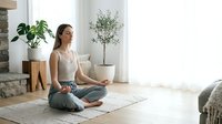 Young woman practicing yoga meditation in bright modern living room with plants and natural light creating peaceful wellness atmosphere at home.