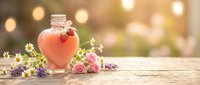 Heart-shaped glass bottle with pink liquid surrounded by wildflowers on wooden surface with warm bokeh lights creating romantic atmosphere.