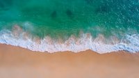 Aerial view of turquoise ocean waves meeting sandy beach shoreline, white foam washing onto pristine sand creates natural pattern in tropical paradise setting.