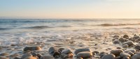 Peaceful pebble beach at golden hour with gentle waves washing over smooth stones creating serene coastal atmosphere for relaxation and meditation.