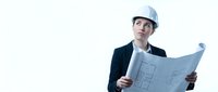 Young male architect or engineer wearing hard hat and dark business suit reviewing blueprint plans against white background.