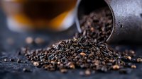 Loose black tea leaves spilling from vintage metal scoop onto dark surface with soft focus background, creating rich texture and aromatic mood for culinary design.
