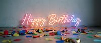 Glowing neon Happy Birthday sign with colorful confetti scattered on the floor, vibrant party decoration against a textured wall.