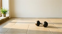 Dumbbell resting on tiled floor in bright, minimalist gym studio with potted green plant by window, ready for strength training workout.