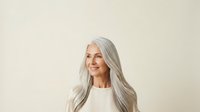 Mature woman with long silver hair and fair complexion wearing white top against neutral beige background, suitable for senior lifestyle, wellness, and beauty campaigns.