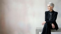 Senior woman with short white hair wearing black blazer and dark clothing, sitting on white bench against neutral background, looking away thoughtfully. Professional portrait for business, corporate, lifestyle, and editorial use.