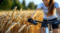 Young woman in casual wear cycling through golden wheat field at sunset, handlebars in focus with ripe grain stalks in foreground creating natural summer atmosphere.