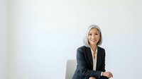 Mature woman with white hair, wearing dark blazer, smiling confidently while seated against white background. Suitable for corporate, business, and professional website design.