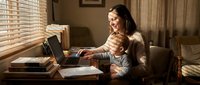 Young mother with toddler son working from home on laptop at wooden desk in cozy living room with warm natural lighting from window blinds.