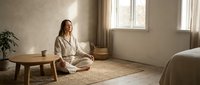 Young woman in white pajamas practicing meditation in minimalist bedroom with natural light streaming through windows creating peaceful morning atmosphere.